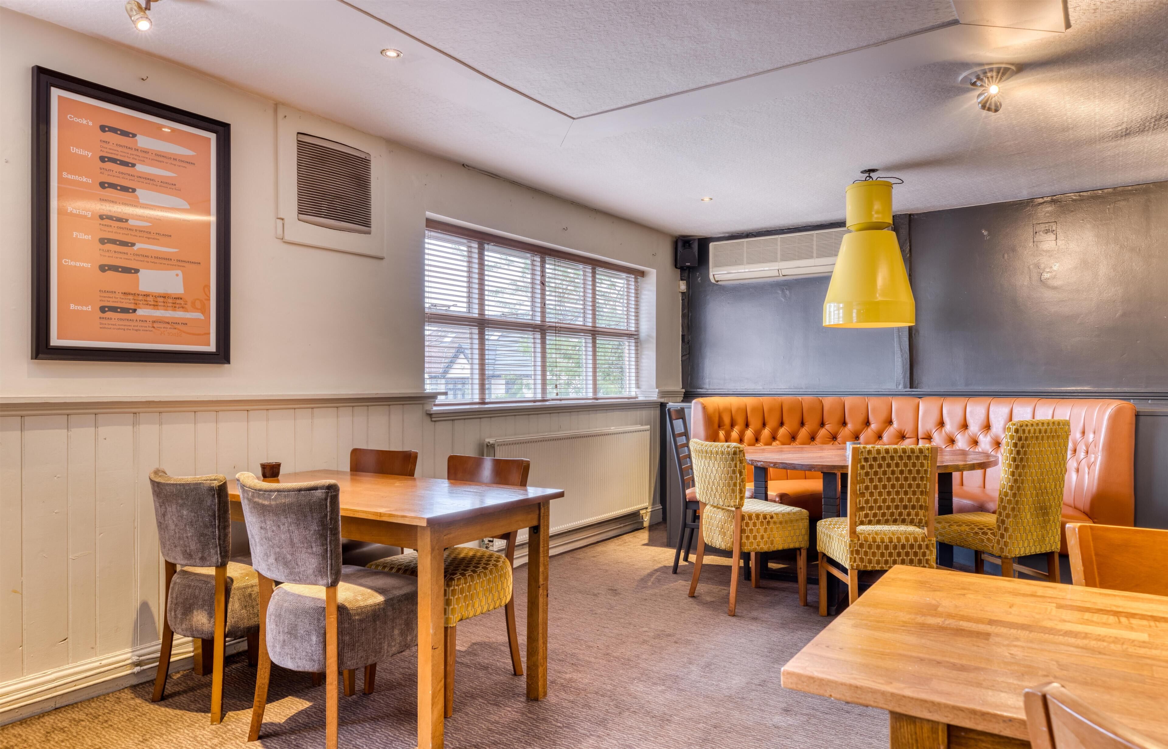 L’intérieur d’un restaurant avec une table et des chaises en bois, un luminaire suspendu jaune, un tableau d’affichage et une fenêtre avec des stores.