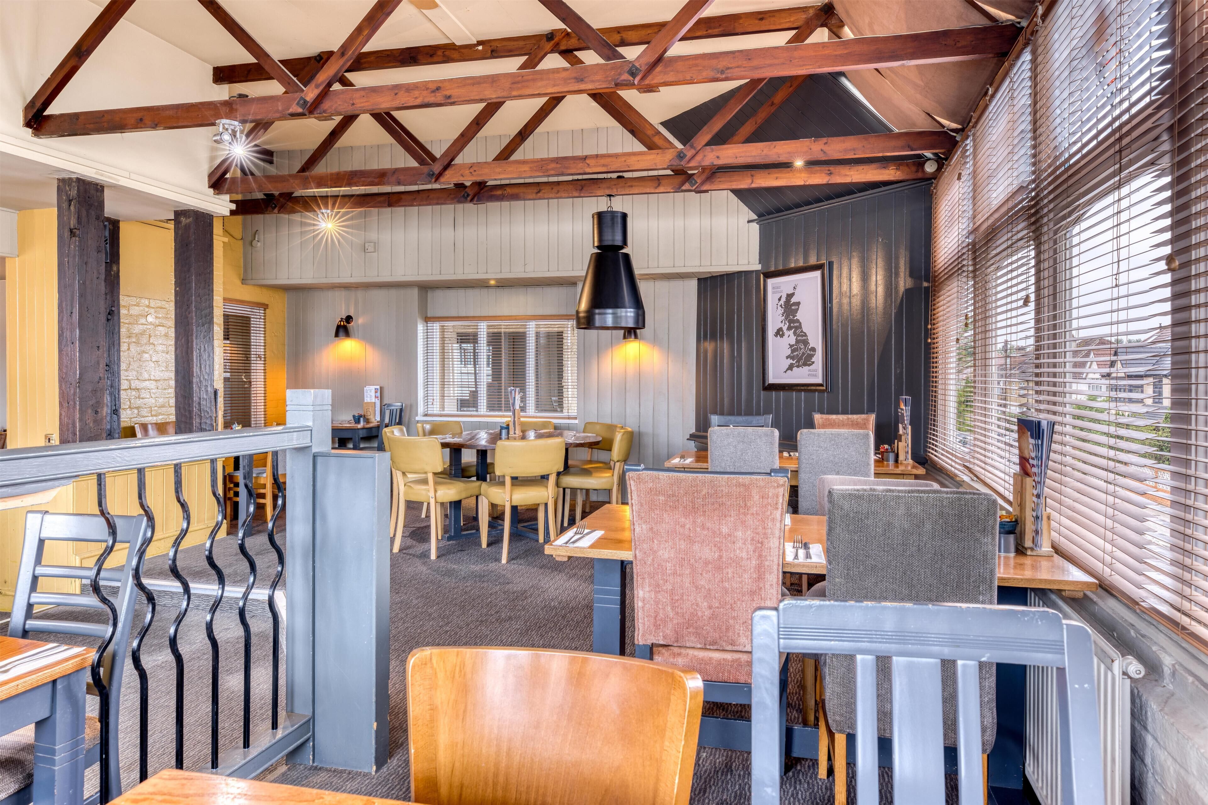 Restaurante com vigas de madeira, mesas, cadeiras e vista através de grandes janelas.