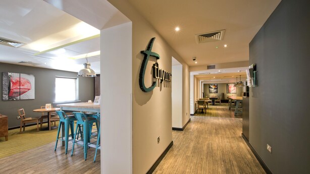 A modern interior with a reception desk, teal chairs, and a hallway leading to a dining area. - Premier Inn London Edgware (Edgware)