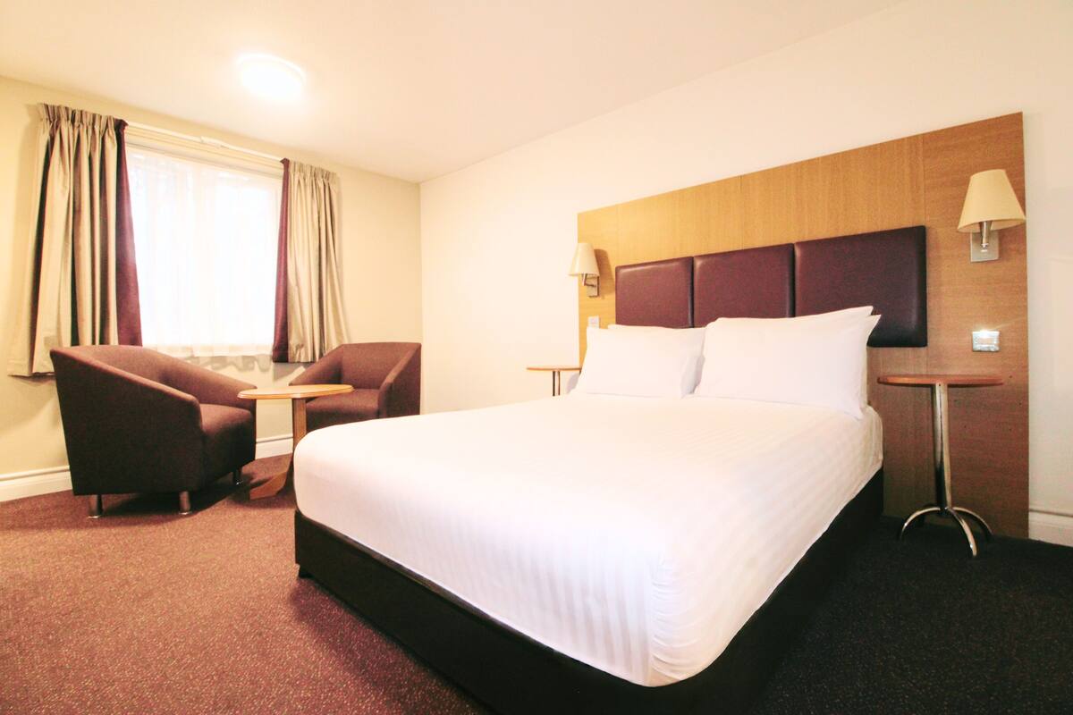 Une chambre d’hôtel avec un grand lit, deux fauteuils, une petite table et une fenêtre avec des rideaux.