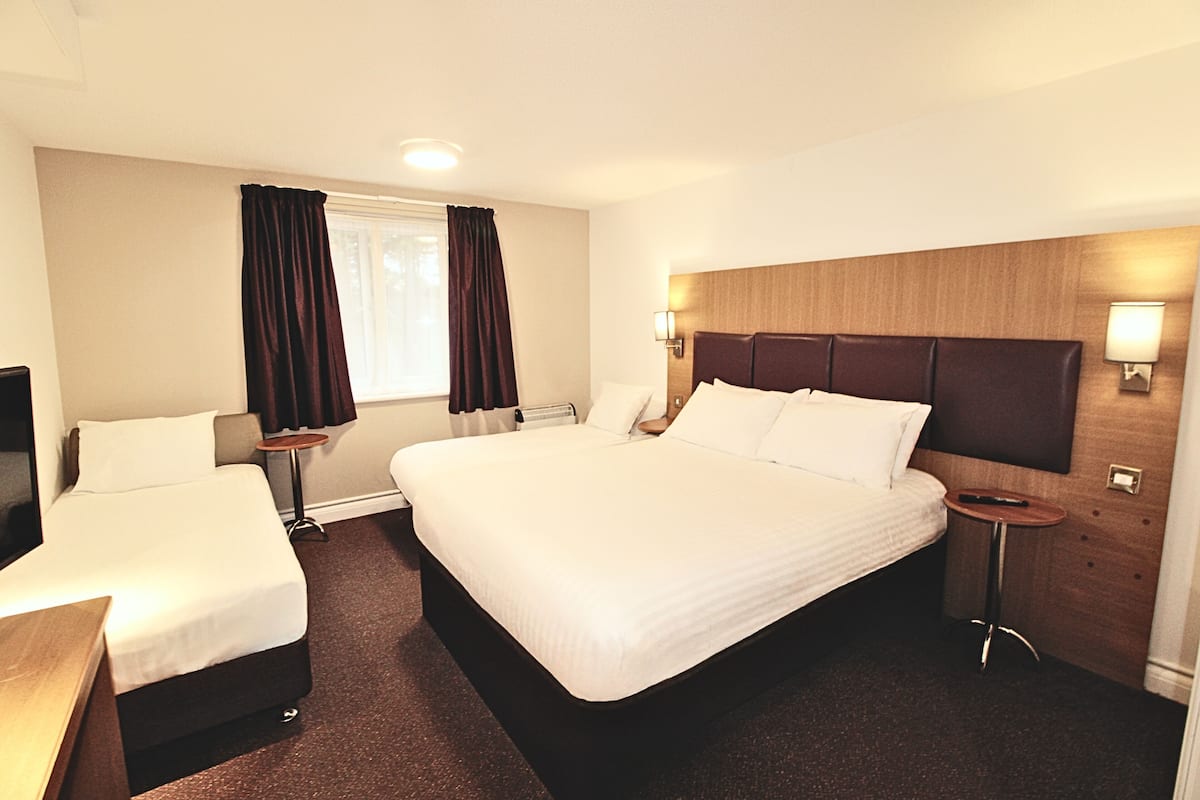 Une chambre d’hôtel avec un grand lit, un lit plus petit, un bureau, une chaise, une fenêtre avec des rideaux et une lampe fixée au mur.