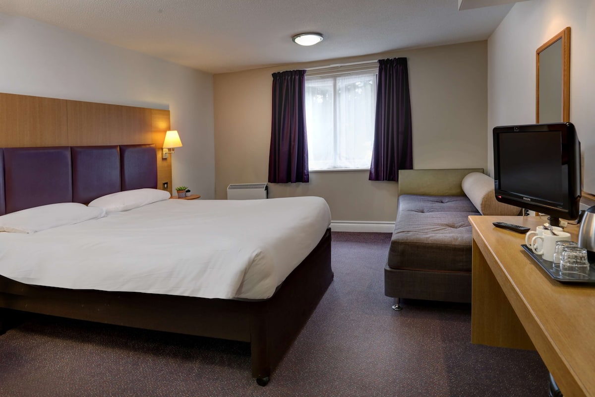 Une chambre d’hôtel avec un grand lit, un bureau avec un ordinateur, une chaise, une petite table avec une lampe et une fenêtre avec des rideaux.
