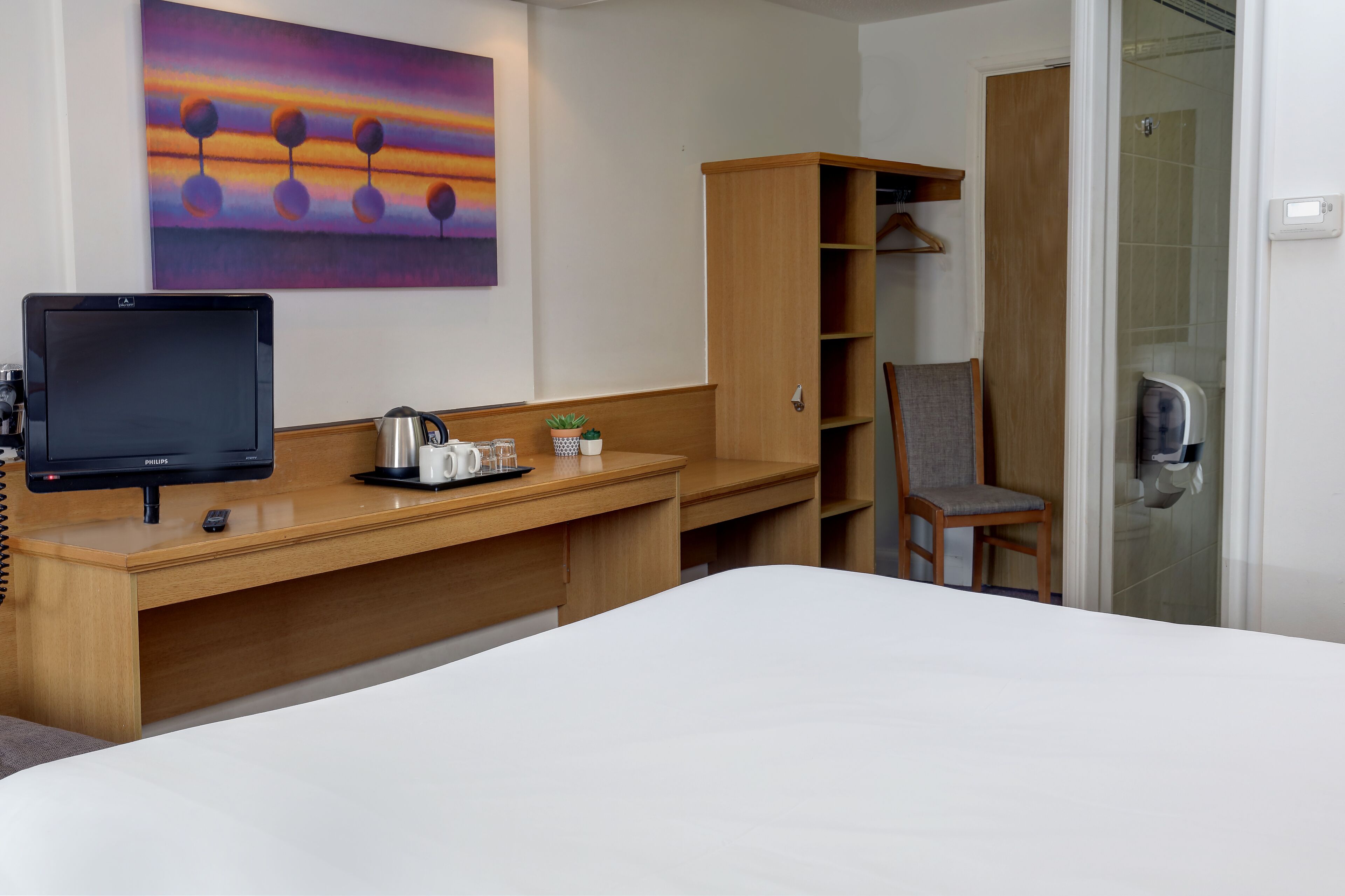 Hotellihuone, jossa on sänky, työpöytä tietokoneella, tuoli, vaatekaappi ja suihku.