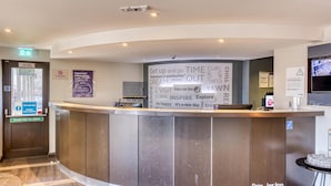 Un mostrador de recepción de hotel con un cartel que dice 'Get up and go TIME OUT' y 'DREAM'. Se ve una señal de salida de incendios, un cartel de prohibido fumar y una barra de empuje para abrir la puerta.