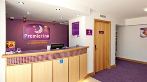 Zona de recepción de hotel con pared morada, mostrador de recepción, puerta de madera con la leyenda 'Privado' y un cartel que indica servicio las 24 horas.