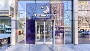Entrada de un hotel con puerta de cristal, un letrero que dice 'Premier Inn' y vista de la calle y edificios al fondo.