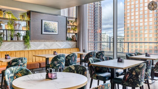 Un restaurante moderno con techo alto, amplios ventanales, una zona de estar con bancos de madera y una variedad de plantas.