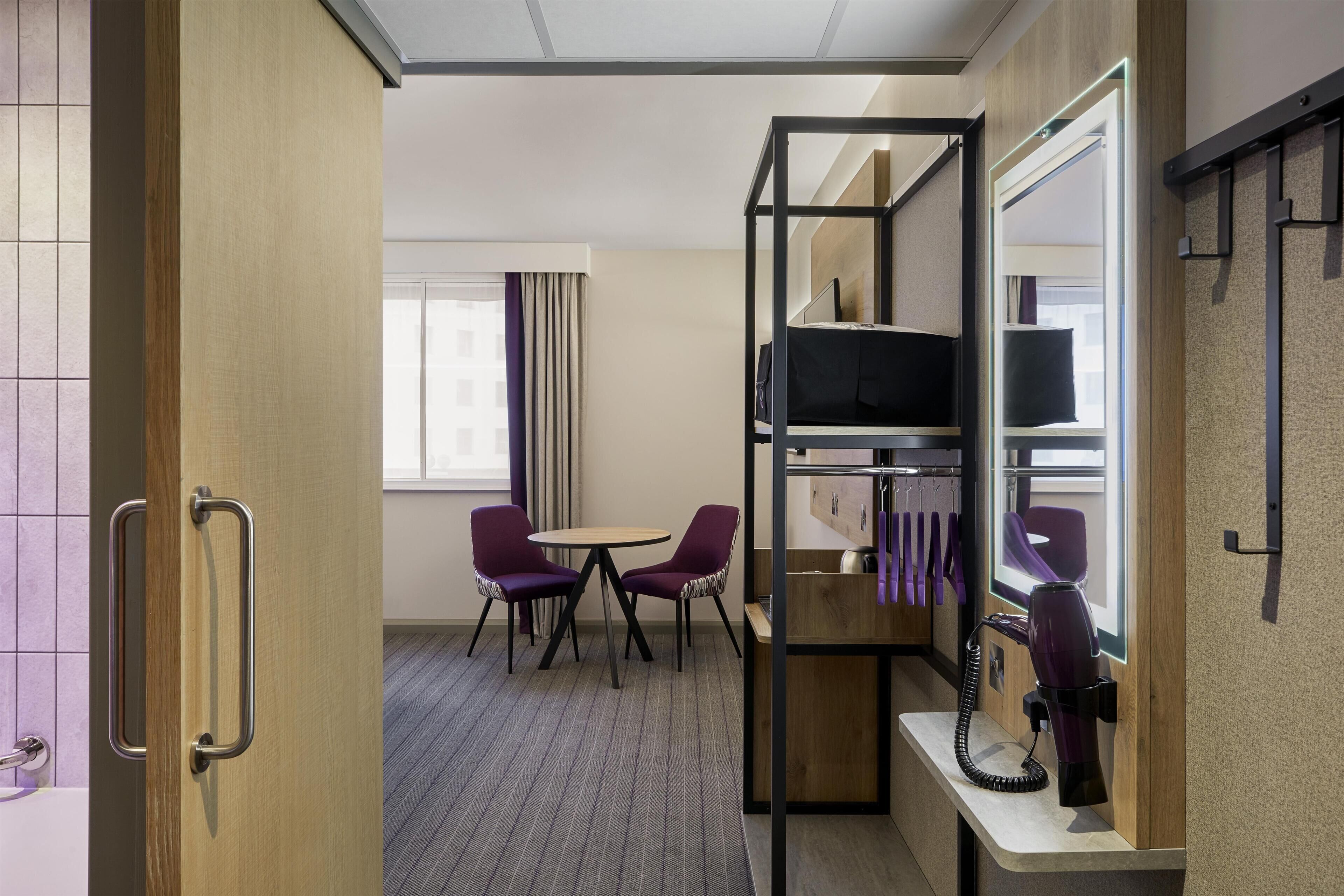 A hotel room with a bed, a TV, a desk with a hairdryer, two purple chairs, and a window with curtains.