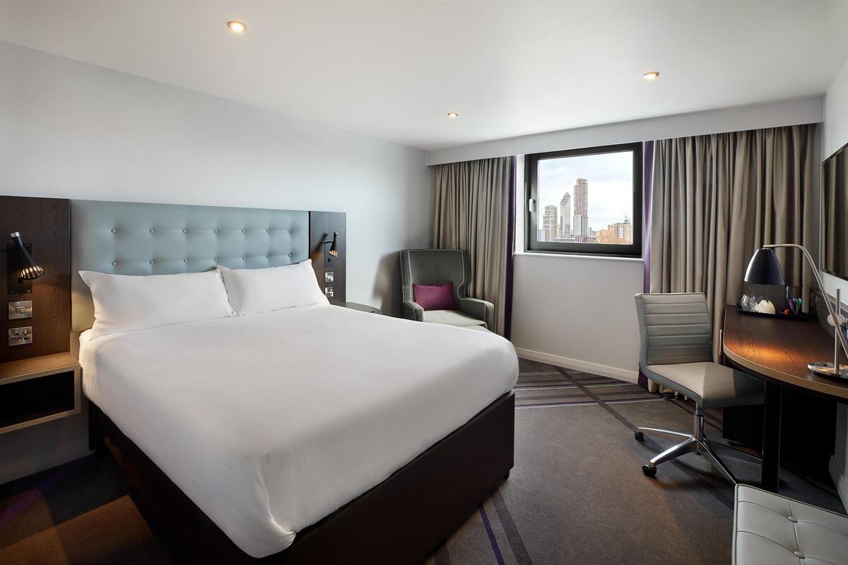 A hotel room with a large bed, a desk, a chair, and a view of the city.