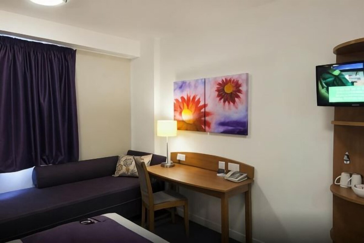 Una habitación de hotel con una cama, un escritorio, una silla, un televisor y un cuadro en la pared.