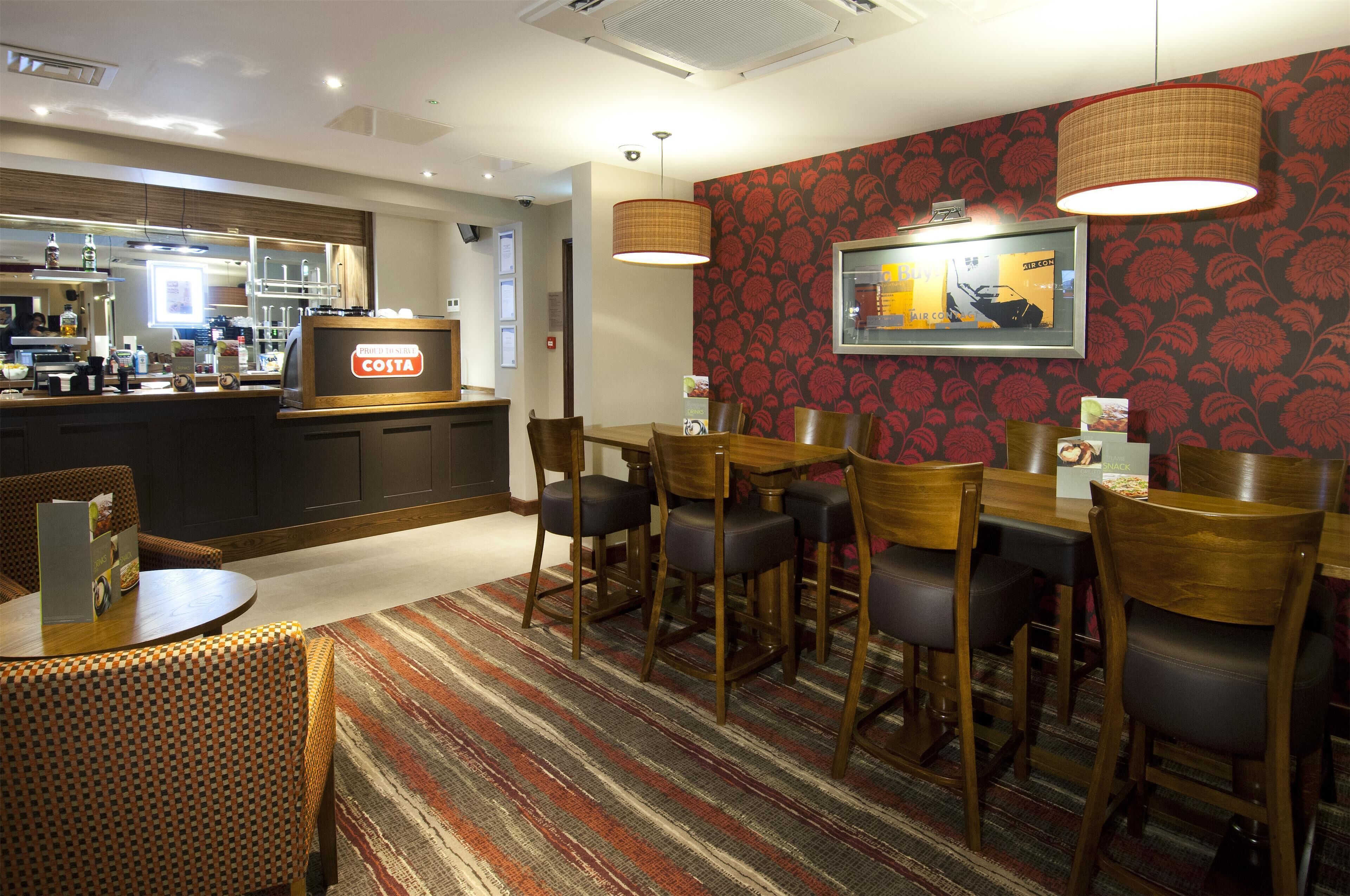 Ein Restaurantbereich eines Hotels mit Bar, Holzhockern, einem gemusterten Teppich und einer Theke mit Costa-Branding.