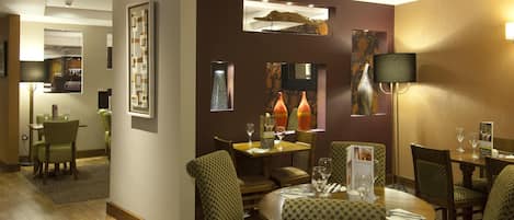 A hotel dining area with patterned chairs, tables set with glasses, and a wall-mounted lamp.