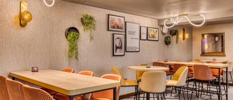 A modern dining area with wooden tables and orange chairs, framed artwork on the walls, and decorative lighting.