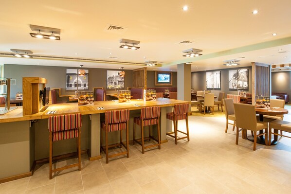 A modern hotel bar with a long counter, striped bar stools, and a television screen. - Premier Inn London Hendon - The Hyde (London)