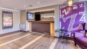 Zona de recepción de hotel con pantalla digital, mostrador de recepción, pared morada con detalles decorativos y un área de descanso con una silla y una mesa pequeña.