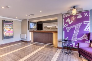 Zona de recepción de hotel con pantalla digital, mostrador de recepción, pared morada con detalles decorativos y un área de descanso con una silla y una mesa pequeña.