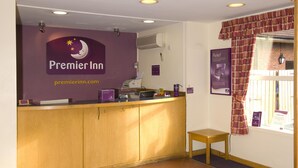 A reception desk with a sign for Premier Inn, a computer, a small table, and a window with curtains. - Premier Inn Liverpool - West Derby (Liverpool)