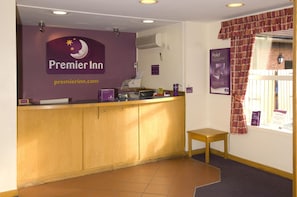 A reception desk with a sign for Premier Inn, a computer, a small table, and a window with curtains. - Premier Inn Liverpool - West Derby (Liverpool)
