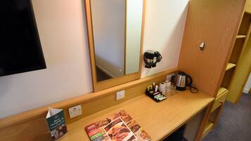 Quarto de hotel com penteadeira de madeira, espelho, suporte para revistas, chaleira e uma pequena prateleira com artigos de higiene pessoal.