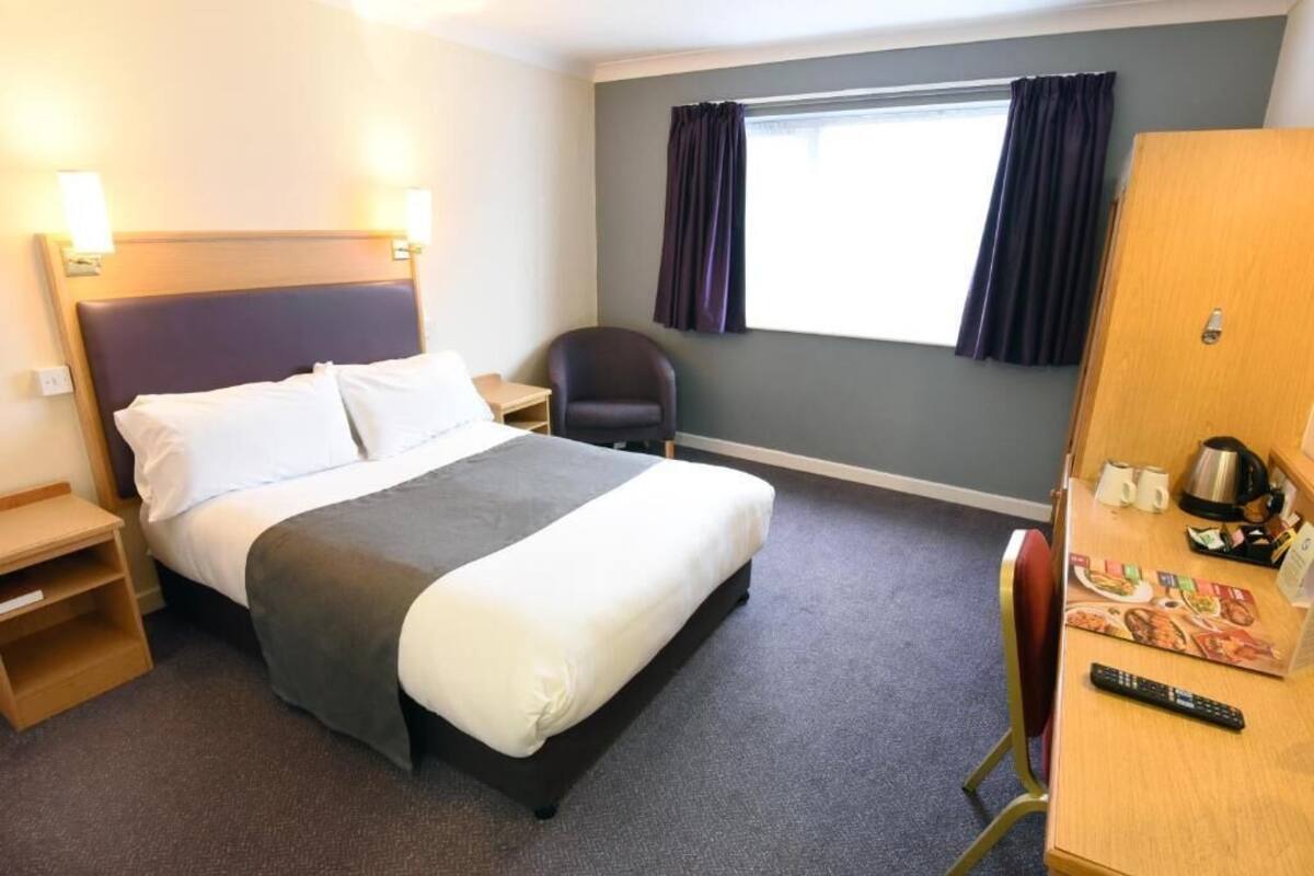 A hotel room with a bed, a desk with a kettle, a chair, and a window with curtains.