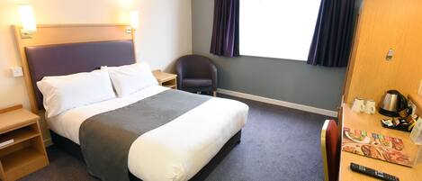 A hotel room with a bed, a desk with a coffee maker, a chair, and a window with curtains.