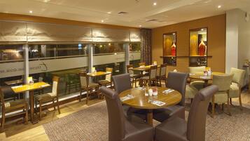 A hotel dining area with multiple tables and chairs, large windows, and a view of the city.