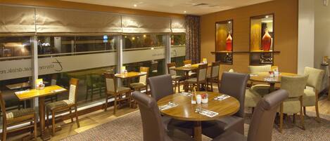 A hotel dining area with multiple tables and chairs, large windows, and a view of the city.