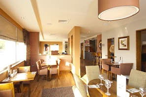 A hotel dining area with multiple tables and chairs, a kitchen in the background, and a view through a window. - Premier Inn Liverpool John Lennon Airport (Liverpool)