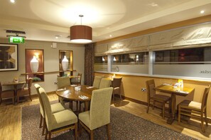 A hotel dining area with a large round table, multiple chairs, a hanging light fixture, and a window with curtains.