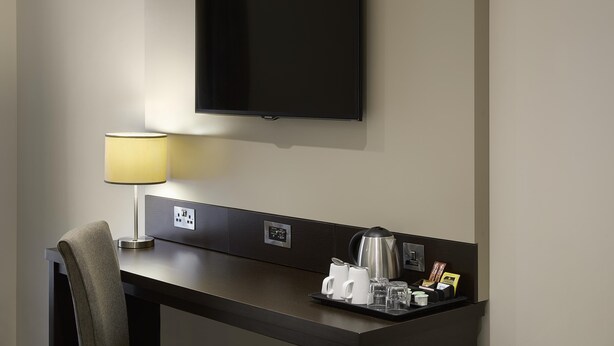 Camera d'albergo con TV a schermo piatto, una scrivania con lampada, una sedia, un vassoio con tazze da caffè e un bollitore, e un pannello di controllo a parete.
