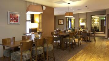 Een restaurantruimte in een hotel met houten tafels en stoelen, hanglampen en kunstwerken in lijsten aan de muren.