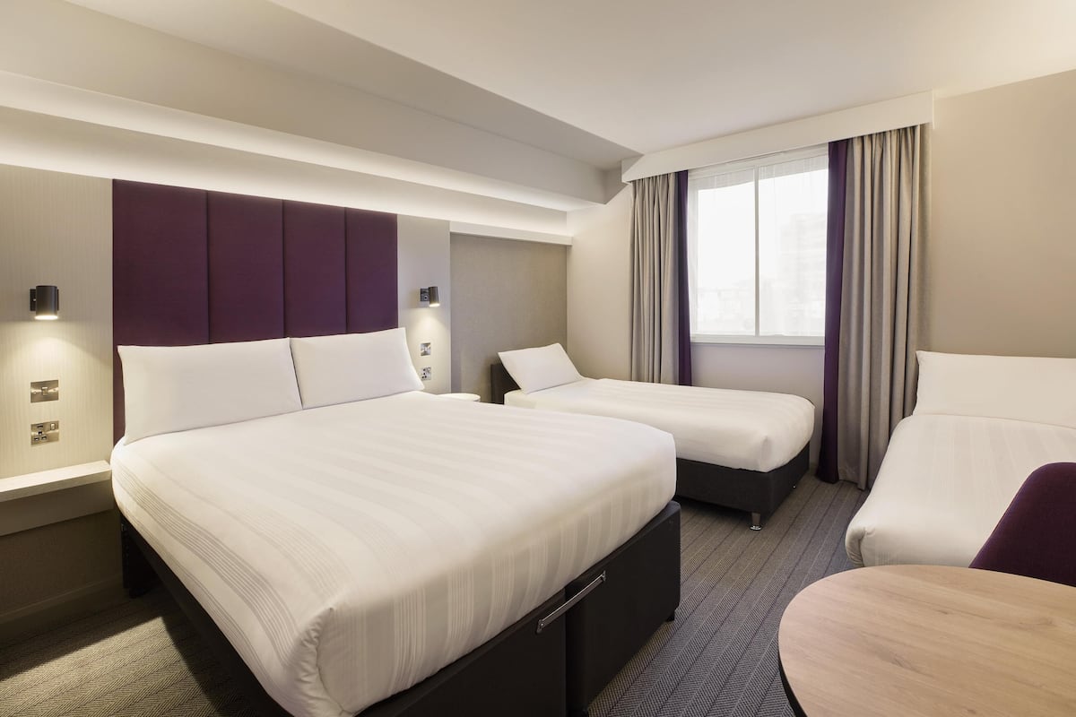 Ein Hotelzimmer mit zwei Betten, einem Schlafsofa, einem kleinen Tisch und einem Fenster mit Vorhängen.