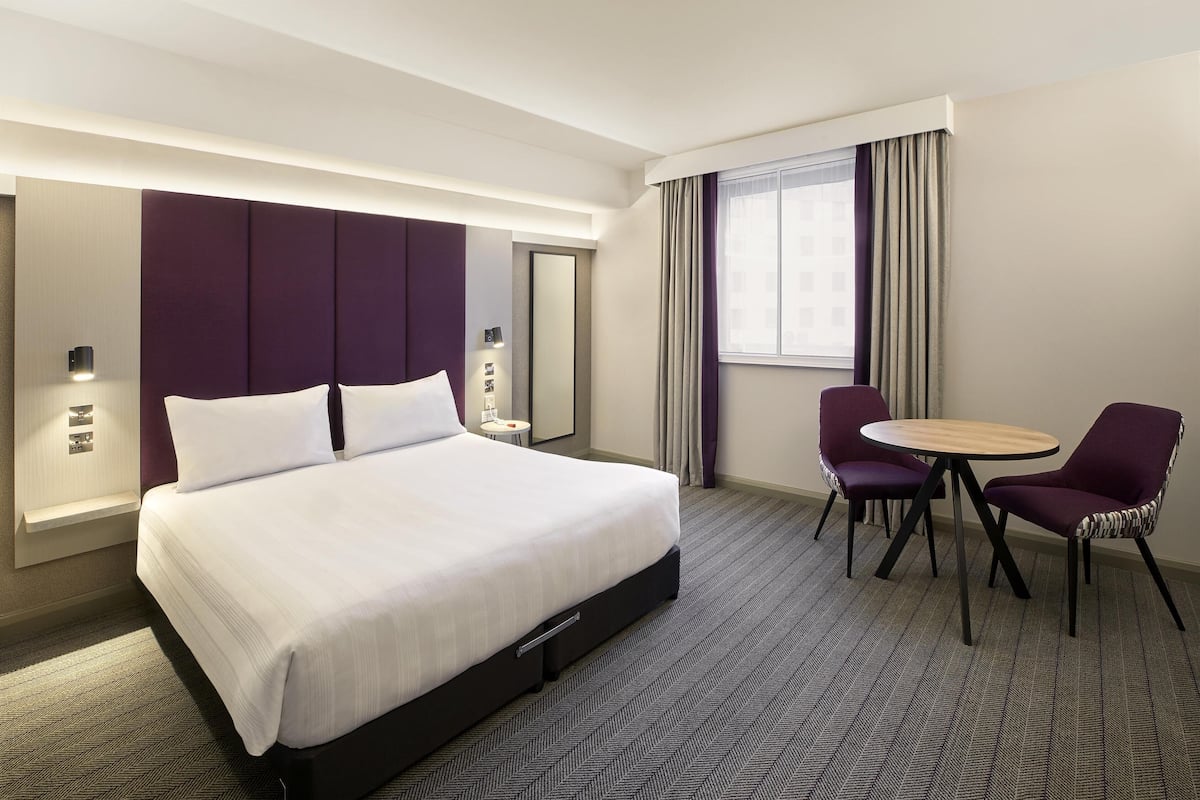 Ein Hotelzimmer mit einem großen Bett, einem runden Tisch, zwei Stühlen, einem Spiegel und einem Fenster mit Vorhängen.