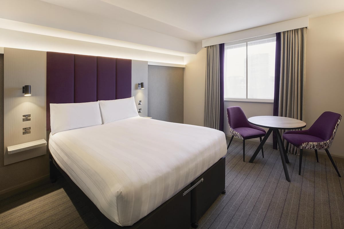 Ein Hotelzimmer mit Bett, Nachttisch, Lampe, Stuhl, kleinem Tisch und einem Fenster mit Vorhängen.