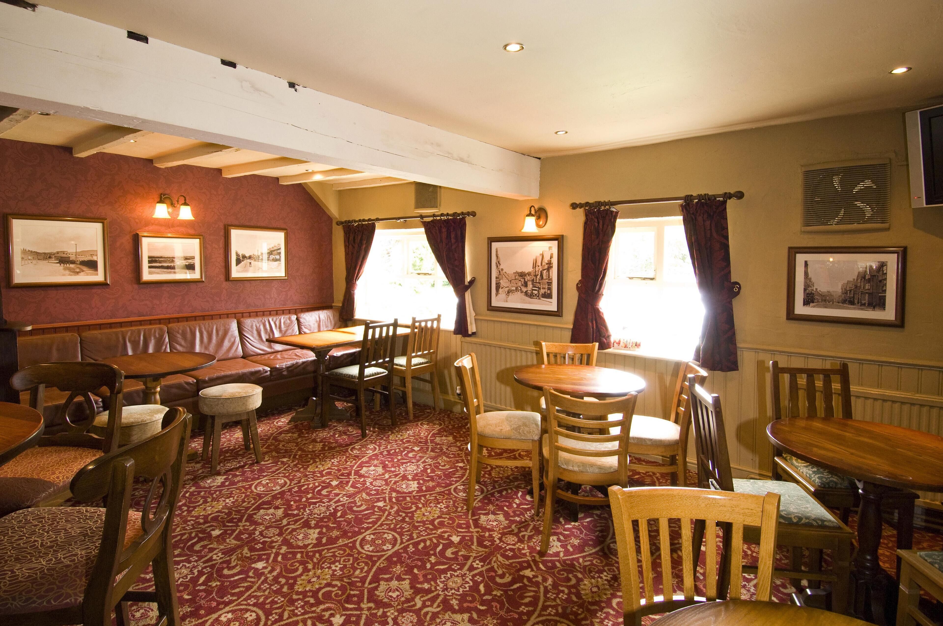 A dining area with wooden tables and chairs, a red patterned carpet, and framed pictures on the walls.