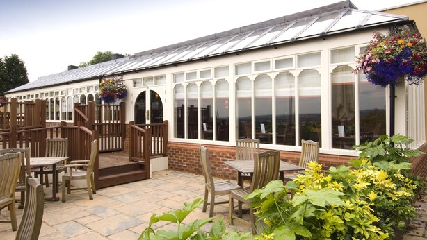 A conservatory with a glass roof, wooden furniture, and hanging flower baskets. - Premier Inn Tring (Tring)
