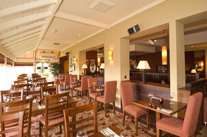 Un comedor de hotel con mesas y sillas de madera, alfombra estampada y techo con vigas de madera.