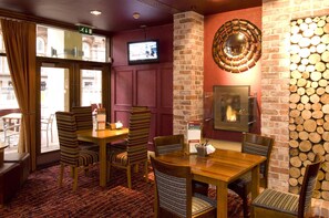 A restaurant interior with wooden tables and striped chairs, a brick wall with a log display, a fireplace, and a television mounted on the wall. - Premier Inn Manchester Central (Manchester)