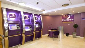 Ein Empfangsbereich eines Premier Inn mit drei Selbstbedienungskiosken, einem Empfangstresen und einer Zimmerpflanze.