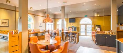A restaurant with round tables, orange chairs, a bar area, and a television screen.