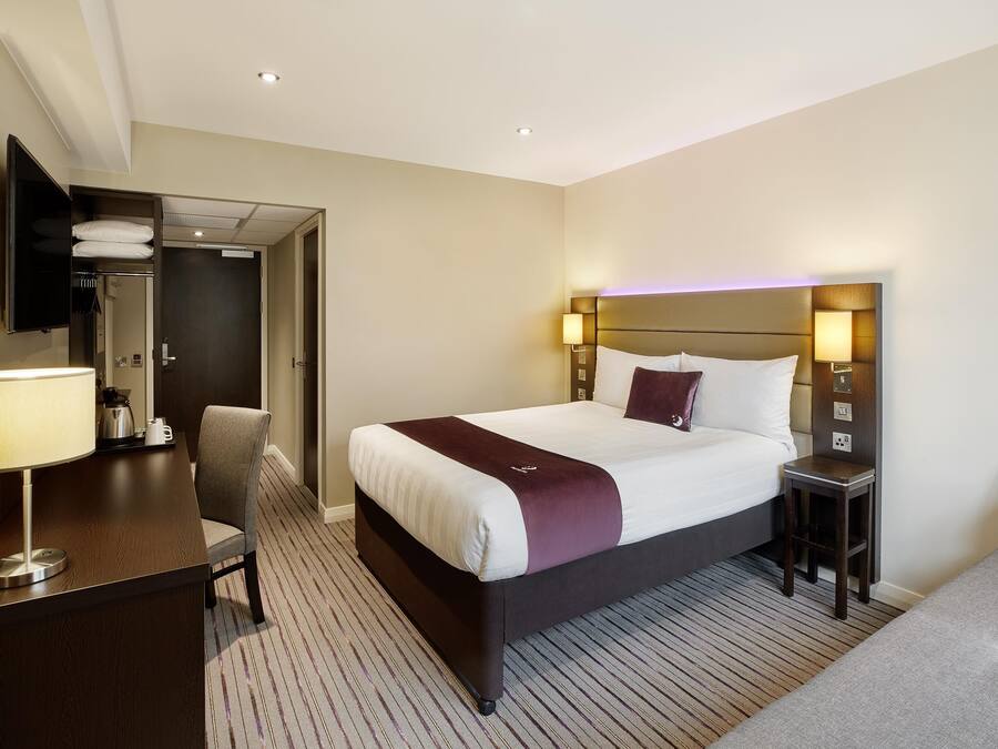 Quarto de hotel com cama, mesa de cabeceira, luminária, cadeira e escrivaninha com luminária.