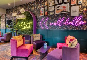 Eine moderne Lounge-Bereich mit komfortablen Sitzgelegenheiten, einem Neon-Schild mit der Aufschrift 'well hello' und einer grünen Wand mit der Aufschrift 'For the most back'.