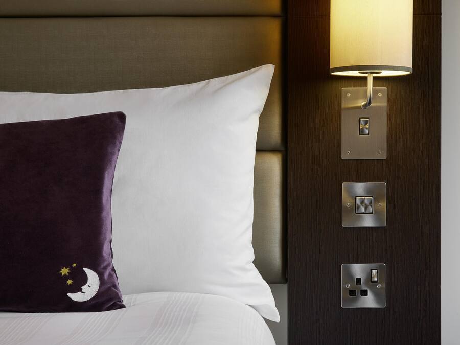 A bed with white and purple pillows, a wall-mounted lamp, and a power outlet.