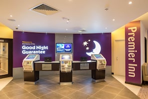 Un espace d’accueil d’hôtel avec un mur violet, un écran affichant des informations et trois bornes d’enregistrement.