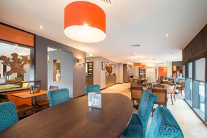 A modern hotel lobby with a round table, blue patterned chairs, a large orange pendant light, and a view of the kitchen area.