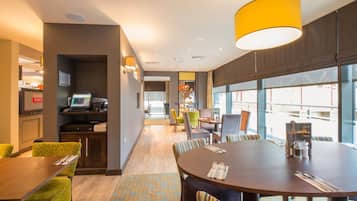 A hotel dining area with a round table, chairs, a yellow pendant light, and a view through large windows.