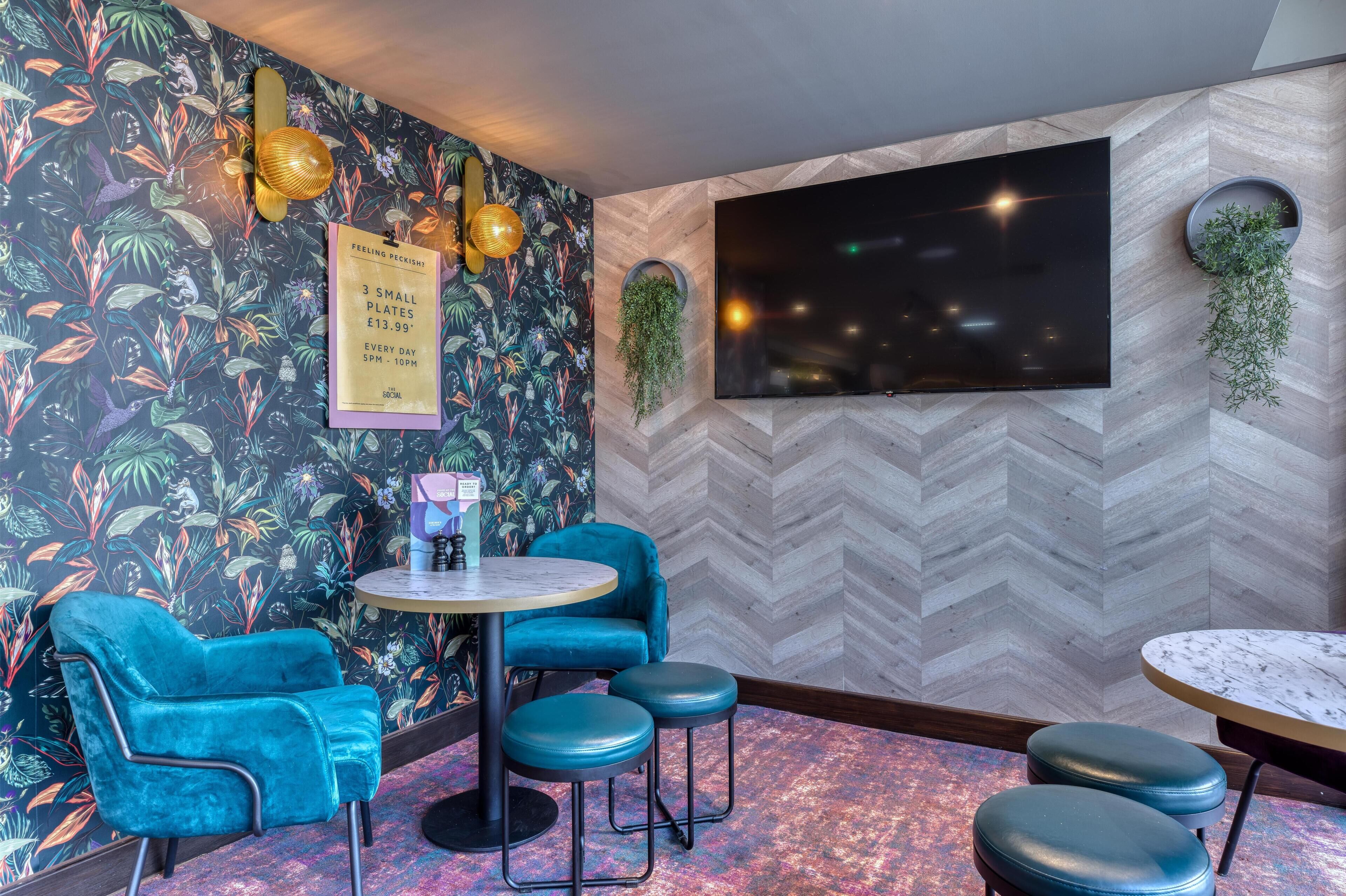 A room with a herringbone-patterned wall, a large flat-screen TV, a round table with stools, a blue velvet chair, and a wall-mounted plant.