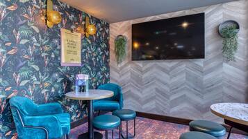 A room with a herringbone-patterned wall, a large flat-screen TV, a round table with stools, a blue velvet chair, and a wall-mounted plant.
