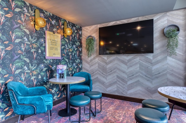 Une pièce avec un mur orné d’un motif en chevrons, un grand téléviseur à écran plat, une table ronde avec des tabourets, un fauteuil en velours bleu et une plante fixée au mur.