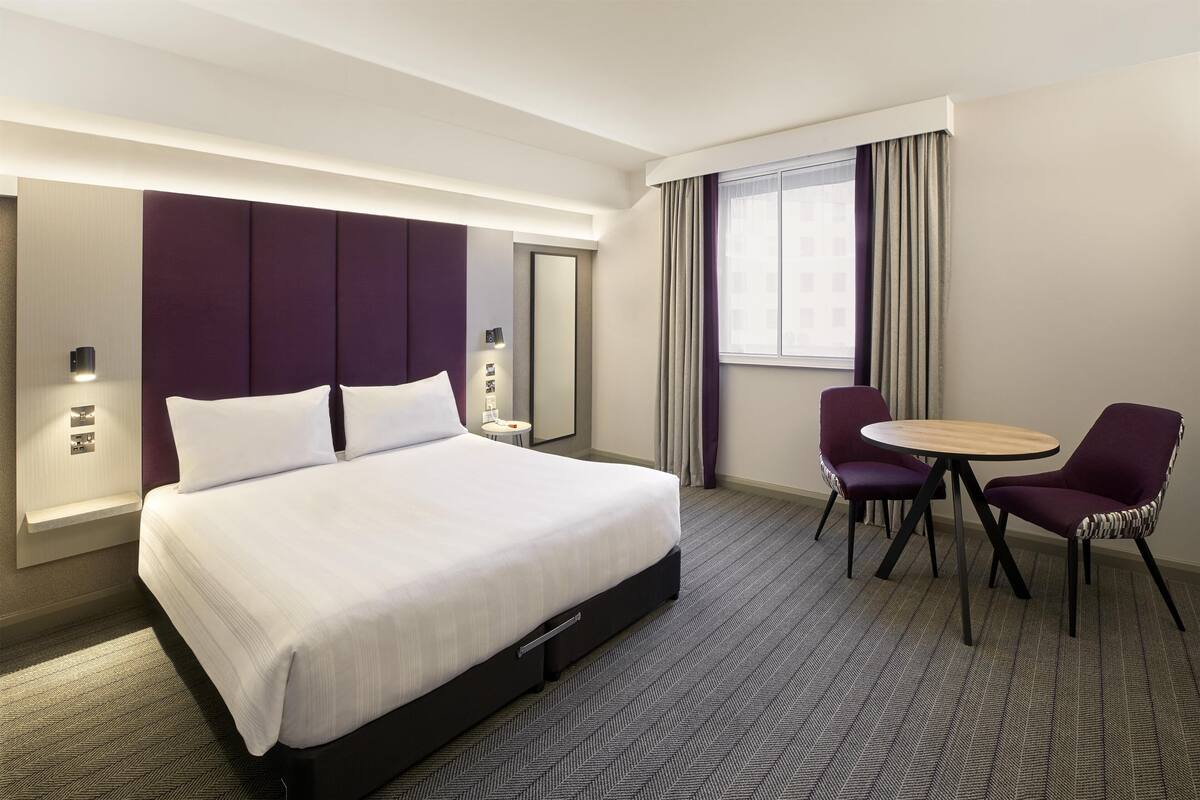 Ett hotellrum med en stor säng, ett runt bord, två stolar, en spegel och ett fönster med gardiner.